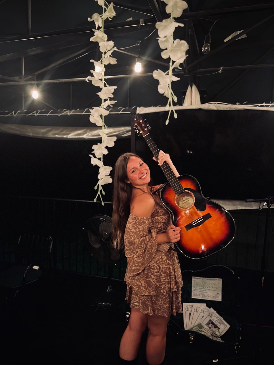 Bella Kollar on a dark stage holding an acoustic guitar beneath hanging white flowers