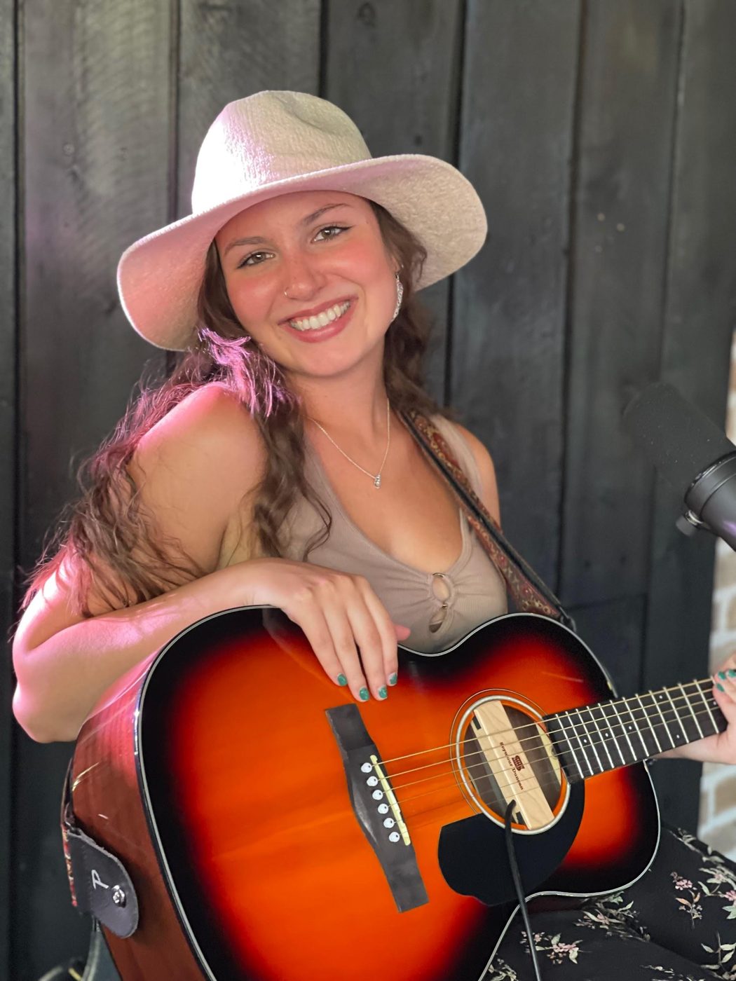 Bella Kollar smiling in a sun hat, holding an acoustic guitar