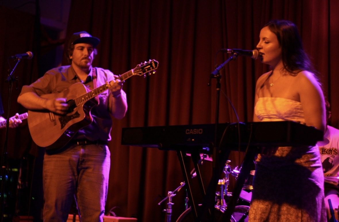 Bella singing at a keyboard beside a guitarist on a red-curtained stage