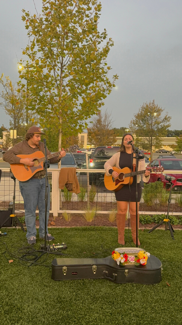 Bella singing and playing acoustic guitar outdoors on the grass beside a guitarist