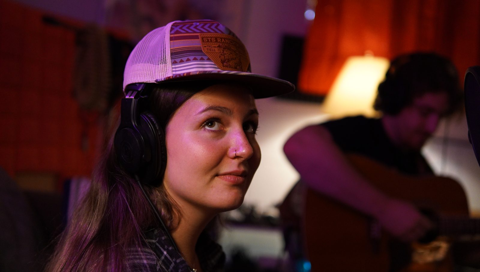 Bella in headphones and a cap listening during a studio session as a guitarist plays