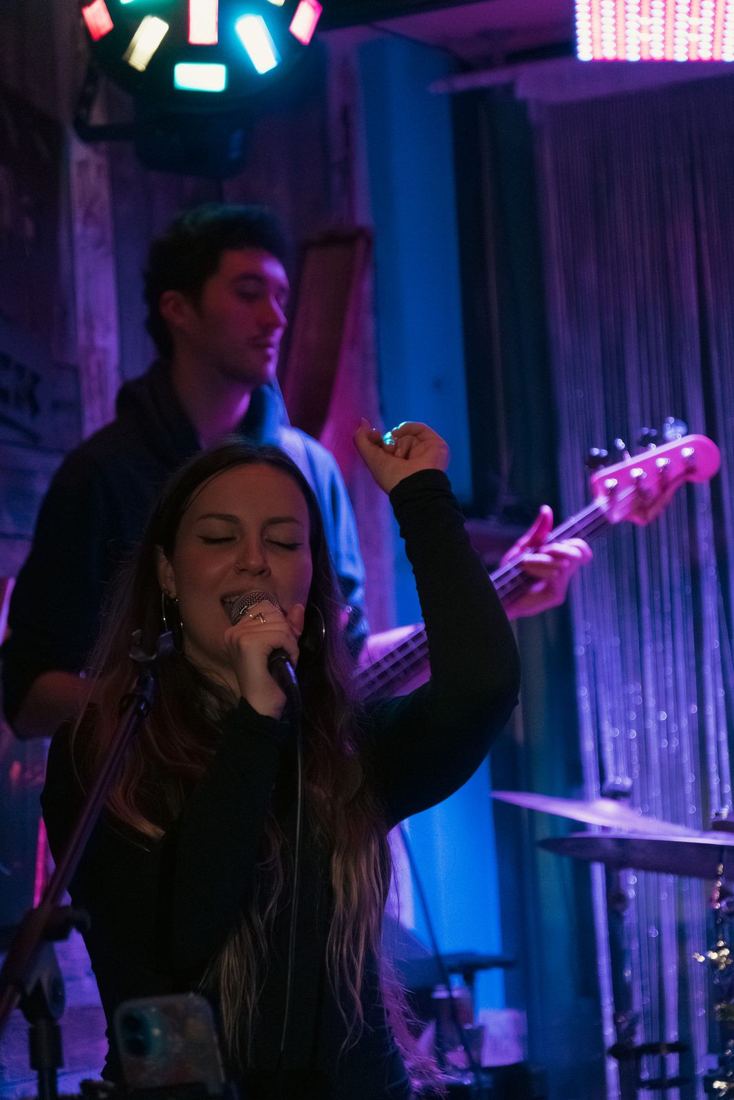 Bella singing into a handheld microphone with one arm raised under blue and purple stage lights